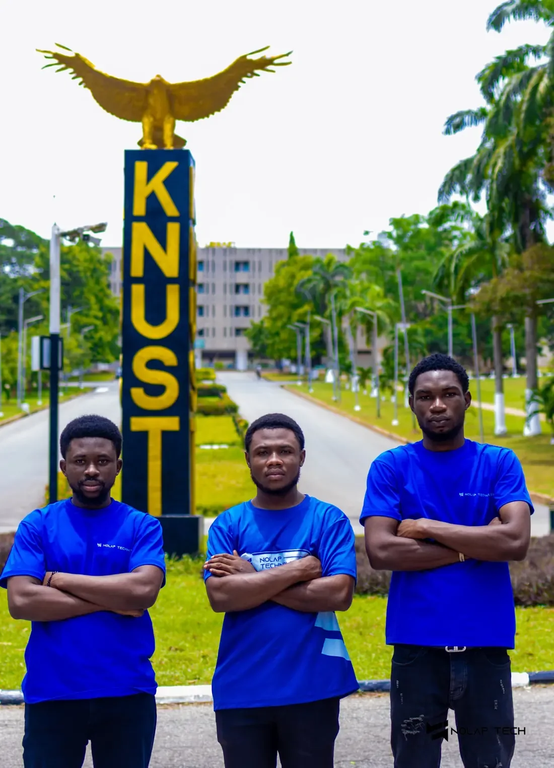 Three NOLAP team members in blue shirts, arms crossed, in front of the KNUST eagle monument